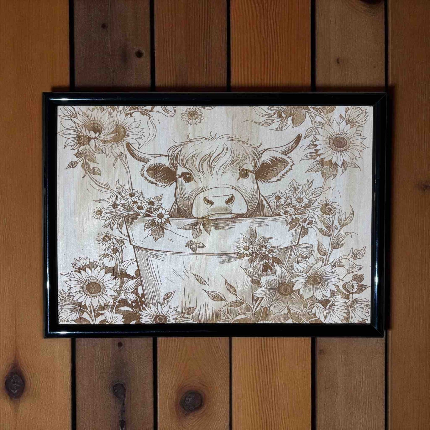 Framed artwork of a cow in a bucket with floral patterns on a wooden wall.