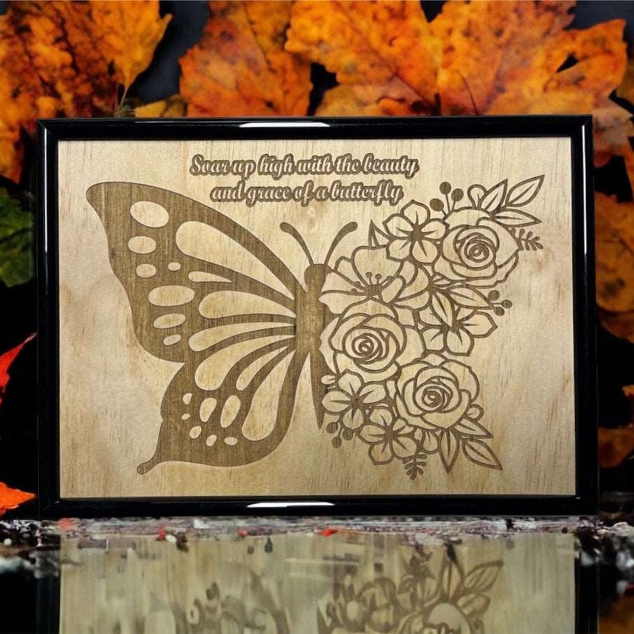 A wooden engraved art piece featuring a butterfly with floral elements and an inspirational quote, displayed against a backdrop of autumn leaves.