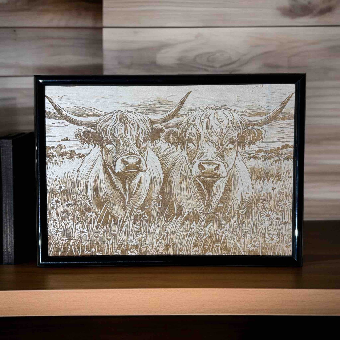 Framed artwork of two highland cows in a field on a wooden surface.