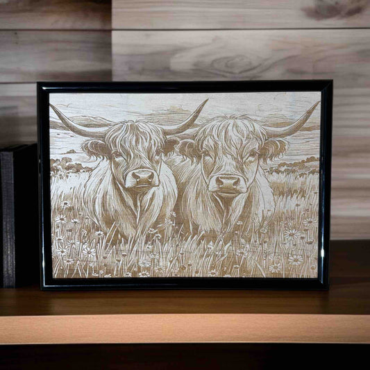 Framed artwork of two highland cows in a field on a wooden surface.