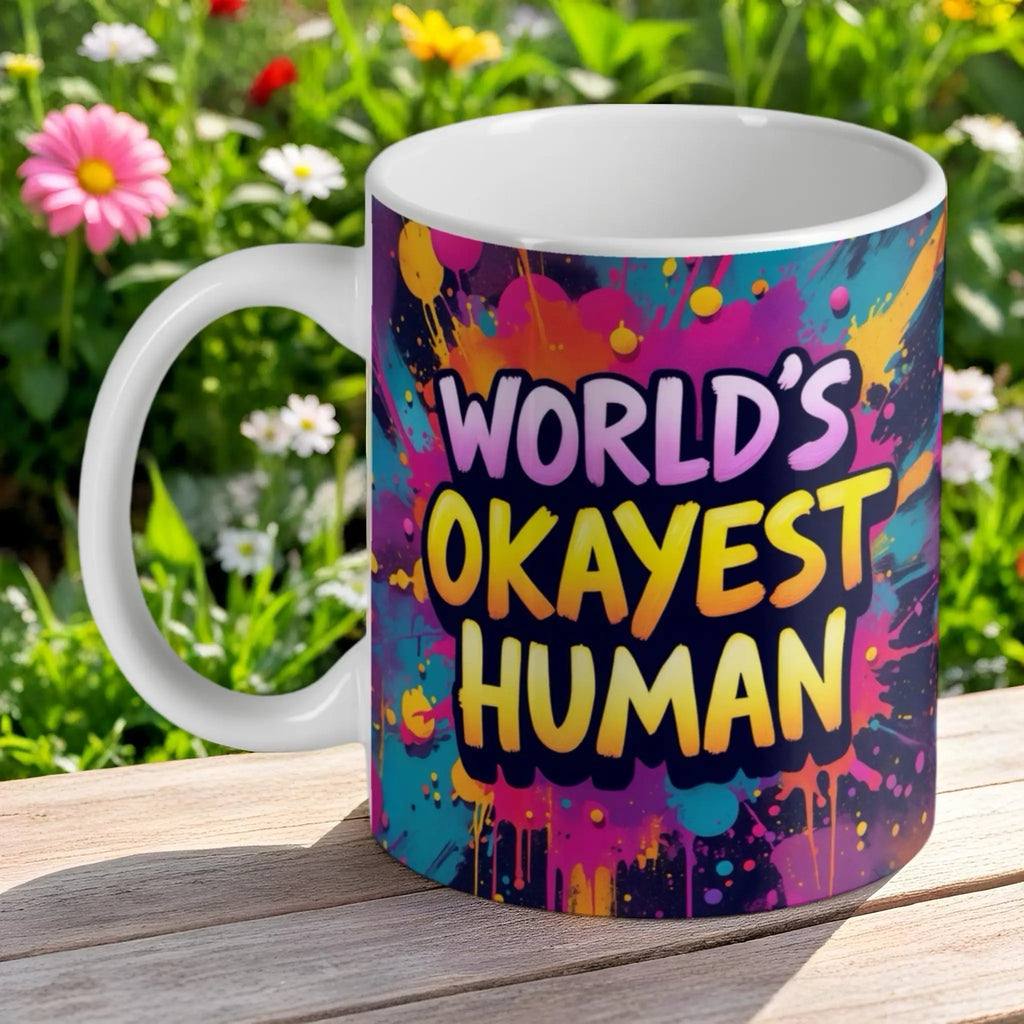 Funny 'World's Okayest Human' ceramic mug by Laser Design Creations, with a colourful graffiti paint splash background.