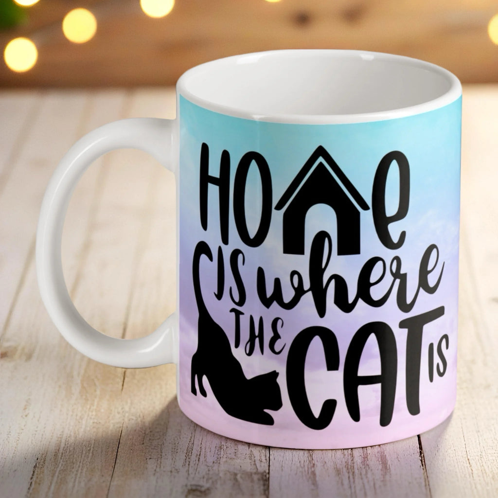 Home Is Where The Cat Is ceramic mug by Laser Design Creations, featuring a cat silhouette against a pastel blue and pink sky.