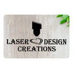 A rectangular sign with a light wood grain texture, featuring the black text 'LASER DESIGN CREATIONS' and a laser pointer graphic. The sign is shown on a white background with a small green leaf in the top right corner.