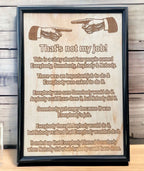Wood engraved framed design with the phrase 'That's not my job!' humorous text, and a gun illustration. Displayed against a wooden background.