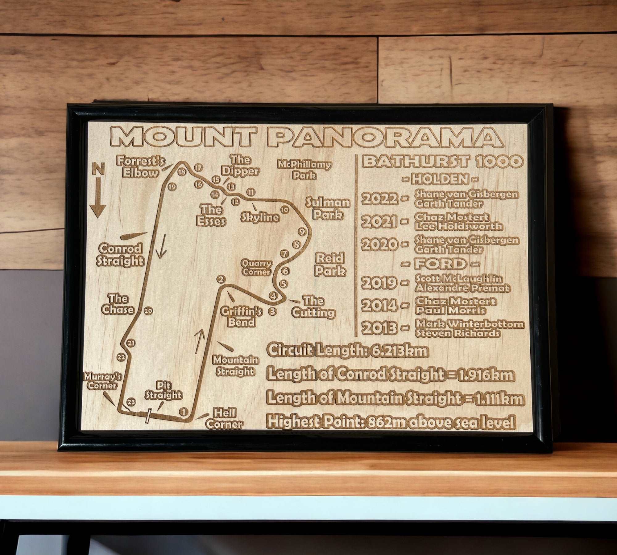 A framed wood engraved wall art depicting the Mount Panorama Circuit, with details of the Bathurst 1000 race, displayed on a wooden shelf.