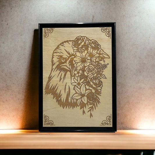 A framed wood engraved artwork depicting an eagle with floral patterns displayed on a wooden surface with a wall in the background.