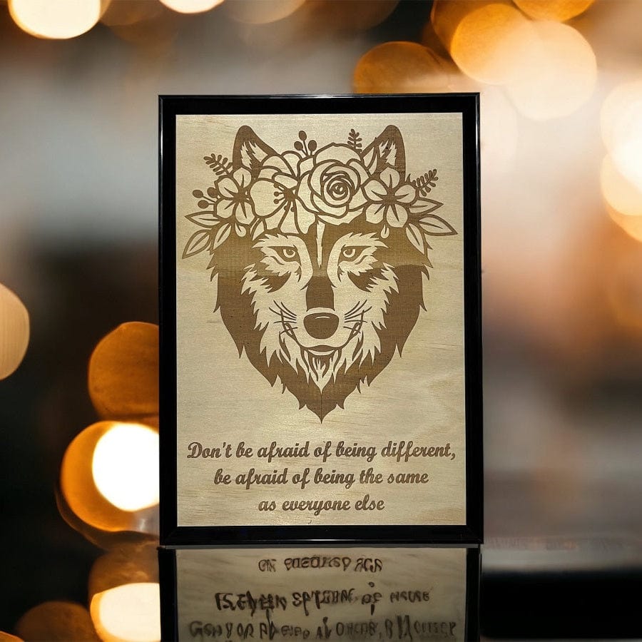 Wood engraved artwork of a wolf with a floral wreath around its head, displayed in a frame with a blurred background.